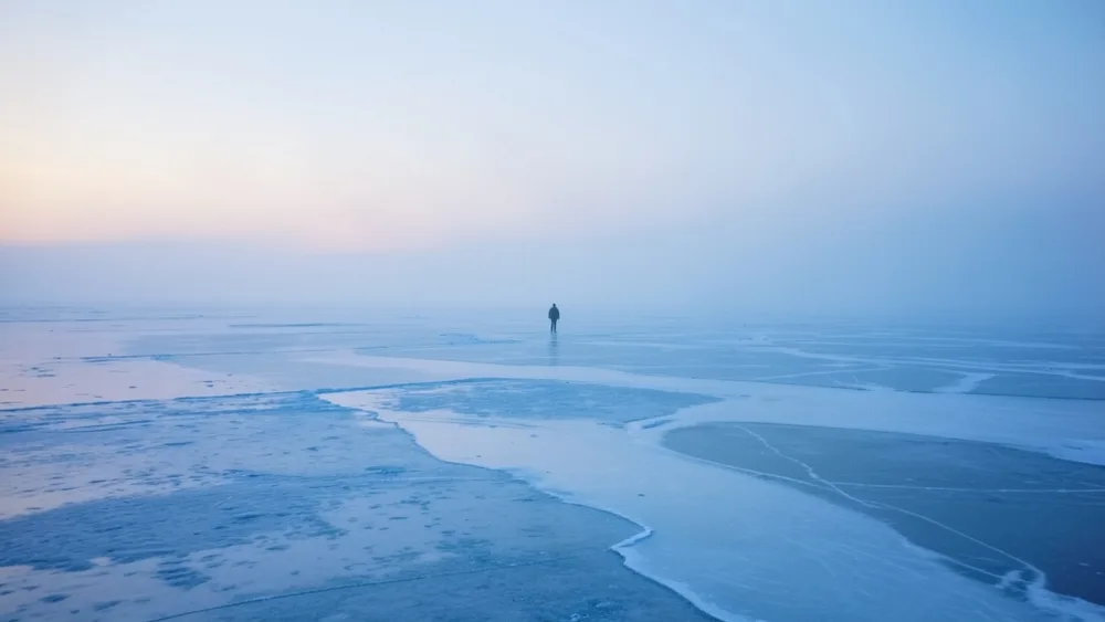 A lone tiny figure walks across a vast pale-blue Arctic ice field in misty dawn light