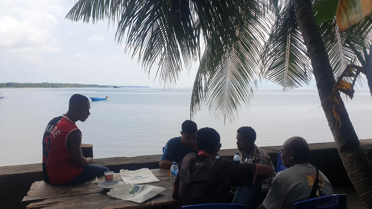 Participants and native speakers sitting at a wooden table on a seaside terrace during a vocabulary recording session in Halmahera