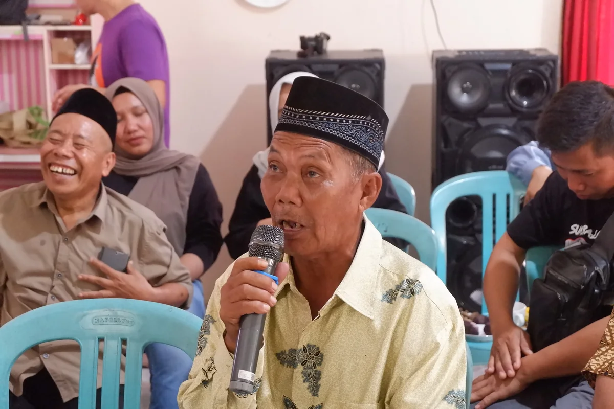 A Balik language speaker in a batik shirt and black cap speaking into a microphone at a community meeting